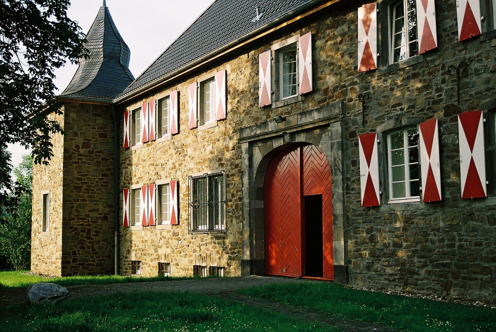 Haus Sülz in Lohmar: natuurstenen gebouw met rood-wit geschilderde luiken en een grote rode poortdeur.